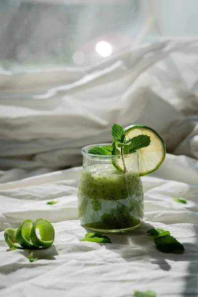 Jar of smoothie with mint leaf. Slice of lime in front of the sun. Green. The lime peel is twisted in a spiral. Top view. On a white cloth with folds. Vertical orientation