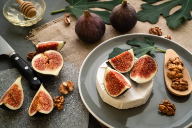 Gri benekli tahtada servis edilir. Gri tabakta incirli camembert peyniri. İncir yaprağı, bıçak, bal ve keten peçete. Yatay yönlendirme.