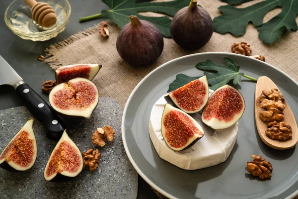 Gri benekli tahtada servis edilir. Gri tabakta incirli camembert peyniri. İncir yaprağı, bıçak, bal ve keten peçete. Yatay yönlendirme.