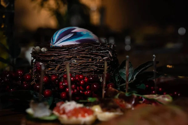 Nest with a blue-white object in the middle. Decorated with red berries. Sandwich with red caviar in the foreground is blurry. Dark tone. Horizontal orientation.