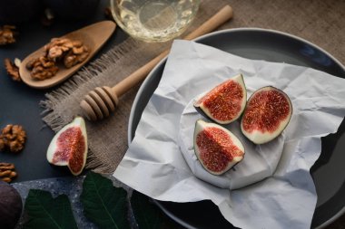 Beyaz kağıdın üzerinde duran yuvarlak peynirli incir dilimleri. Ceviz ve bal için ahşap kaşık yakınlarda. Keten bir masa örtüsünün üzerinde. Koyu gri bir arka planda. Yatay yönelim.
