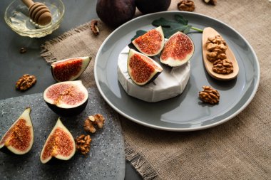 Yanında incir ağacı ve ceviz yaprağı olan gri bir tabakta, camembert peyniri üzerine incir dilimleri. Çarşaf peçeteye. Arkasında tahta kaşık olan bal. Yatay yönelim.