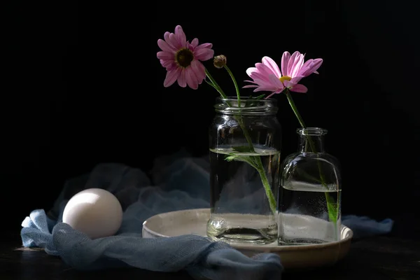 Still life with delicate flowers of daisies with rose petals on a dark wooden background with a kitchen gauze towel and chicken eggs. Easter composition.