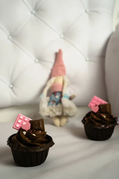 Kahverengi kapkekler açık renkli deri koltukta pembe çikolatayla süslenir ve arka planda da bir tekstil bebeği bulunur..