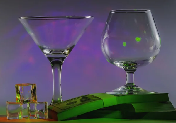 Martini Glass together with Cognac Glass on a packs of the 100s of us dollars and ice cubes with flashing smooth violet lights on background — Stock Photo, Image