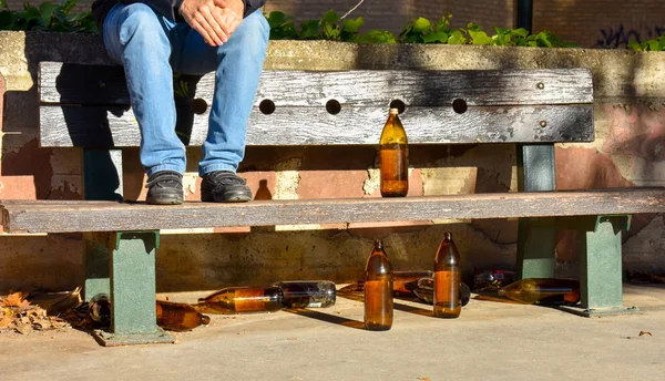 O homem sentou-se em um banco com muitas garrafas laranjas grandes da cerveja feita do vidro completamente vazio no parque devido a alguém bebeu o tempo antes de deixá-los sobre e sob um banco marrom — Fotografia de Stock