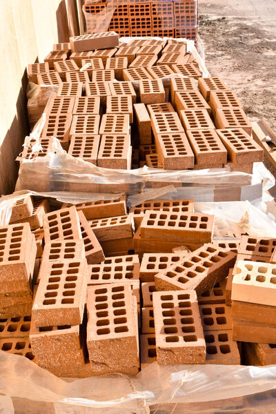 a wooden pallet plenty of old stacked red bricks in rows. Behind there is other pile of red bricks wrapped with plastic.