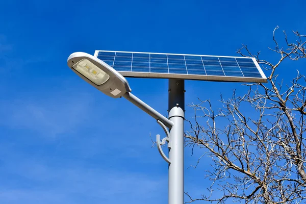 metallic post in a street with a solar panel to generate renewable photovoltaic electricity. The solar panel produces electrical power to light a led bulb in the same post. Vertical