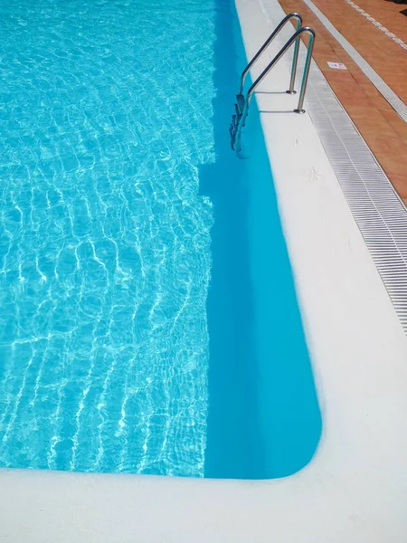 Angolo di una piscina di un luogo turistico pieno di acqua limpida e trasparente. Il bordo della piscina è di colore bianco e ci sono alcune piastrelle marroni intorno e una scala metallica. Verticale — Foto Stock