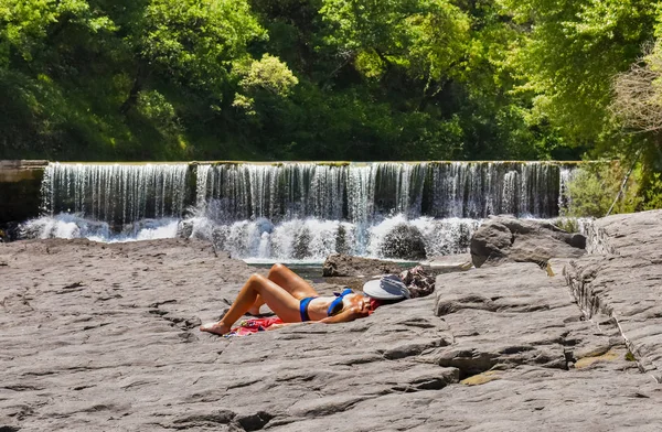 Femme bronzée en maillot de bain est couché sur des rochers gris ayant un bain de soleil et de protéger sa tête avec un chapeau. Sur le fond une cascade d'une rivière de montagne et quelques arbres d'une forêt verte . — Photo