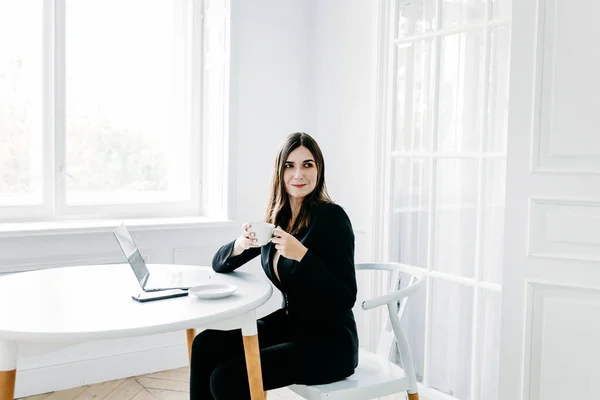 Girl in dark clothes, suit working on a laptop, lawyer, consultant, specialist, work in the office and from home, Internet, communication with colleagues, coffee break, tea, Cup, water, snack