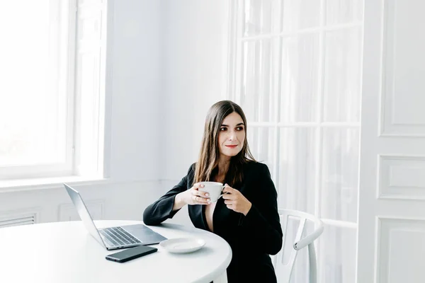 Girl in dark clothes, suit working on a laptop, lawyer, consultant, specialist, work in the office and from home, Internet, communication with colleagues, coffee break, tea, Cup, water, snack