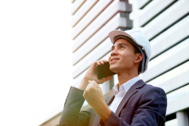 Erkek mühendis mobilephone arka plan gündoğumu bina kullanarak beyaz kask giyiyor.