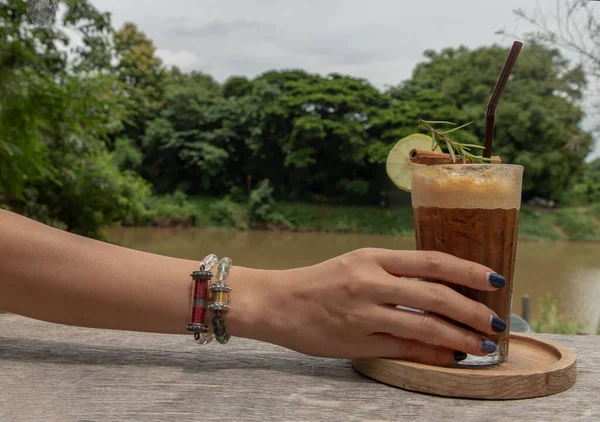 女性的手拿着冰柠檬咖啡 一杯混合蜂蜜和柠檬的咖啡 还有河景 更新夏季饮料的概念 有选择的重点 — 图库照片