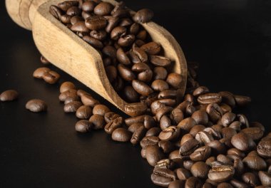 Koyu arkaplanda, ahşap kahve kepçesinde zengin dokulu bir yüzey üzerinde kavrulmuş kahve çekirdekleri manzarası. Yaşamak için sabah kahvenizin tadını çıkarın, onu metin, tasarım, duvar kağıdı ve poster için alan olarak kullanın, seçici odak noktası olarak kullanın.