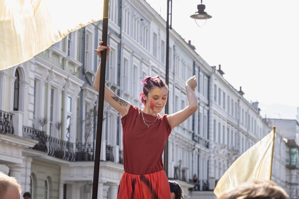 Caught mid-dance, a woman in a striking red outfit holds a flag standard, embodying the vibrant energy of the Notting Hill Carnival. The bright sun illuminates the elegant white buildings in the background, setting the scene for a day of celebration.