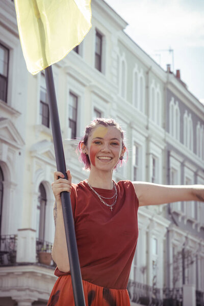 Caught mid-dance, a woman in a striking red outfit holds a flag standard, embodying the vibrant energy of the Notting Hill Carnival. The bright sun illuminates the elegant white buildings in the background, setting the scene for a day of celebration.