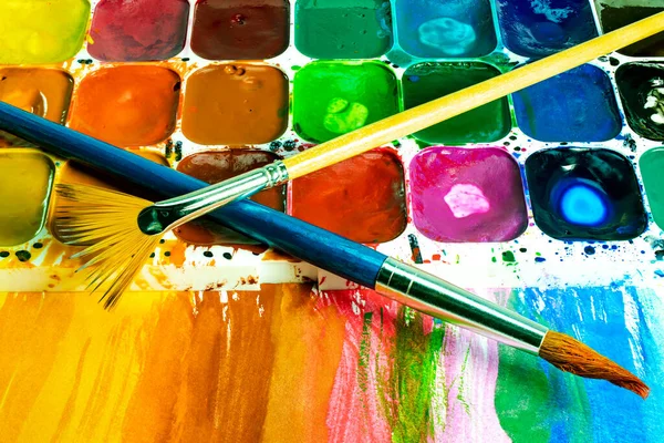 Multi-colored watercolor paints and brushes. Materials for drawing and creativity. Bright water-based paints. School supplies. Hobbies and creativity.