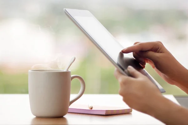 Tablet close up photo with woman hand. Girl using tablet in the cafe and drinking hot coffee. Woman using tablet computer laptop and make note on book with pencil with window glass background — Stock fotografie