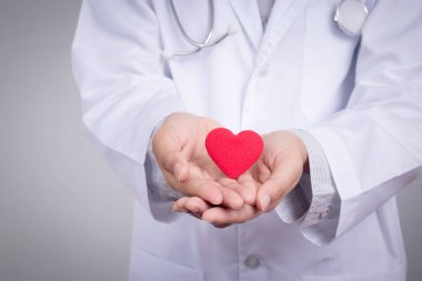 Kırmızı bir kalp taşıyan genç bir kadın doktor, gri arka planda duruyor.