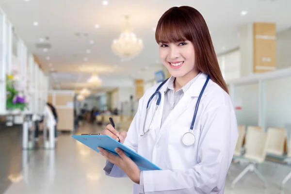 Detail seorang dokter wanita Asia muda memegang clipboard berdiri di atas latar belakang rumah sakit. konsep layanan kesehatan — Stok Foto