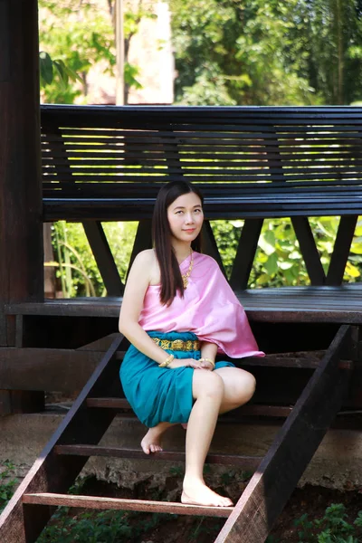 Thai Asian polite woman with smile face in traditional costume of Thailand at waterfront pavilion front of riverside ancient house near the river. Beautiful Asia female model in her 20s.