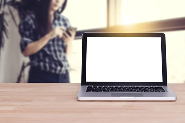 Büroarbeitsplatz mit leerem weißen Bildschirm-Laptop mit Clipping-Pfad auf Holztisch. Frau benutzt Mobiltelefon durch Berührung im Hintergrund in der Nähe des Fensters. asiatisches Frauenmodell — Stockfoto