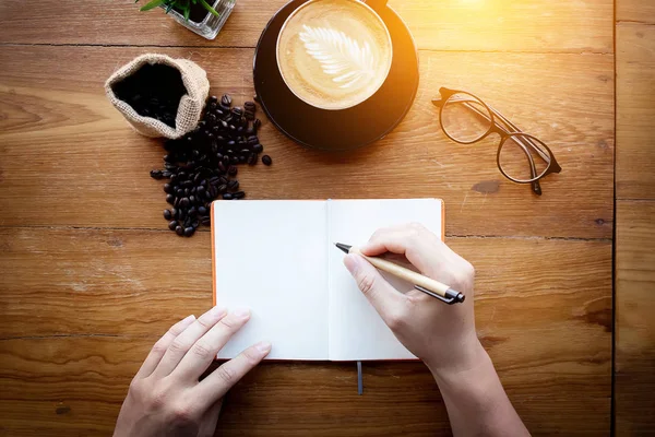 Barista kafe kahve hazırlama hizmeti vermektedir. Cappuccino kahve ve kahve çekirdeği ile vintage ofis masası dekorasyon metin için alan ile boş beyaz sayfada bir kalem yazma notu tutan Adam, üst görünümü — Stok fotoğraf