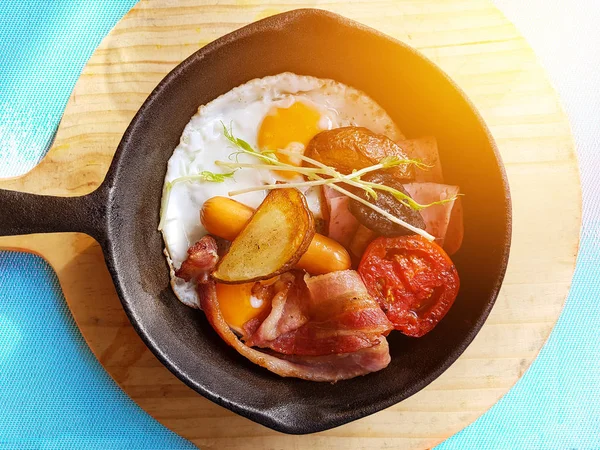 Breakfast in cooking pan with fried eggs, sausages, bacon , tomato , potato and vegetable on wood block background on the table in the kitchen. morning light effect