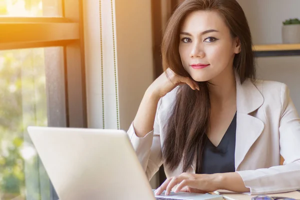 Concepto de startup en Asia. enfocado joven mujer de negocios asiática con cara de pensar trabajando con el ordenador portátil en el lugar de trabajo mirando a la cámara, efecto de película y el sol efecto de destello . —  Fotos de Stock