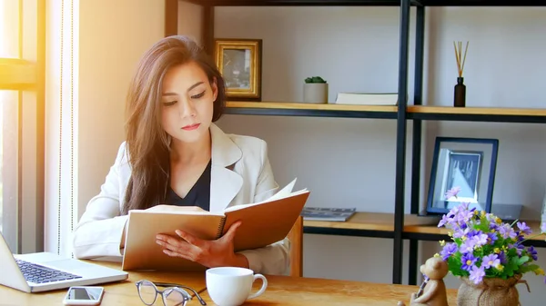 Concepto de startup en Asia. joven mujer de negocios asiática leyendo libro con cara de pensamiento trabajando con el ordenador portátil en el lugar de trabajo, efecto de película y el sol efecto de destello . —  Fotos de Stock