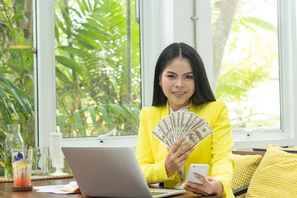 Portret wesoły młody Asian kobieta trzyma banknoty pieniędzy i telefon komórkowy z uśmiechem twarz przed notebookiem w miejscu pracy — Zdjęcie stockowe