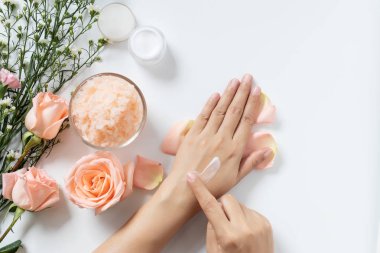 doğal cilt bakımı konsepti. kadın kozmetik krem kavanoz ile beyaz arka plan üzerinde ellerine beyaz krem uygulamak, tuz spa scrub, kopya alanı ile gül ve beyaz çiçekler