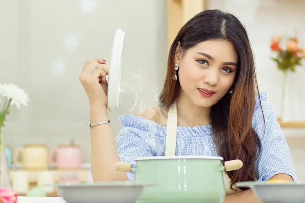 Mooie Aziatische vrouw zitten aan de tafel in de keuken, open de deksel van de soep pot met de warmte damp zwevend omhoog. — Stockfoto