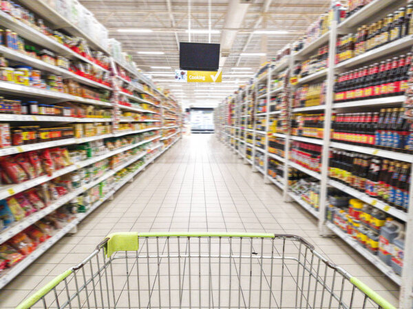 shopping cart with abstract blurred supermarket aisle with colorful goods on shelves at background, sign of product for cooking hanging from ceiling