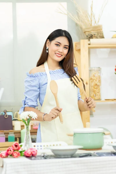 Hermosa mujer asiática sosteniendo cubiertos de madera detrás de la mesa de comedor con olla, plato y utensilios de cocina en la sala de cocina con la cara sonriente — Foto de Stock