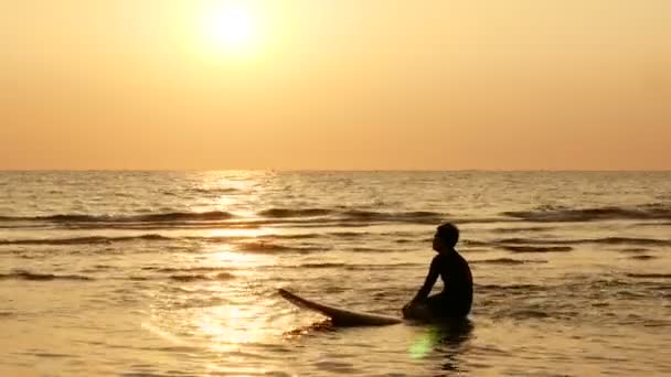 熱帯のビーチで夕日に海の上にサーフボードに座っているサーファーの男のシルエット — ストック動画