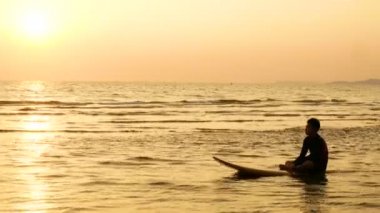 4k. sörfçü adam siluettropikal plajda gün batımında deniz üzerinde sörf tahtası üzerinde oturarak rahatlayın. yaz sezonu konsepti için spor ve rekreasyon