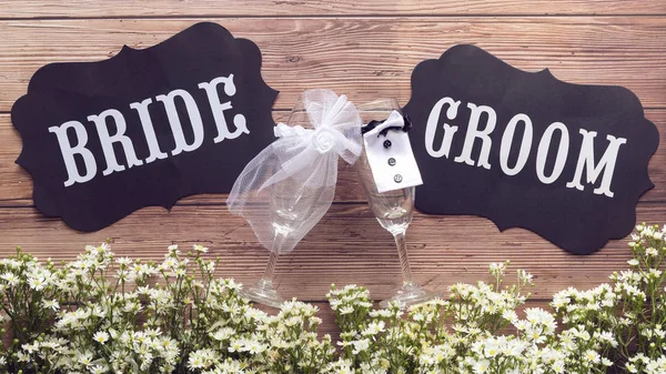 Sklenici šampaňského ve svatebních šatech s textovým znaménkem nevěsta a ženicha na dřevěné pozadí vyzdobené droboučkým bílým květináčem, starobylý styl. koncept svatební známky — Stock fotografie
