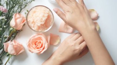 4k. doğal cilt bakımı konsepti. kadın kozmetik krem kavanoz ile beyaz arka plan üzerinde ellerine beyaz krem uygulamak, tuz spa scrub, kopya alanı ile gül ve beyaz çiçekler