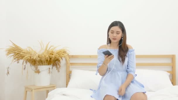 美丽的亚洲妇女坐在床上听音乐与耳机从手机应用和跳舞与放松和快乐的情感 — 图库视频影像