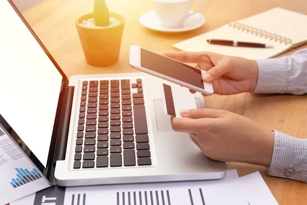 Mujer que sostiene una tarjeta de crédito y compra haciendo el pago en línea a través de teléfono inteligente móvil con pantalla en blanco delante de la computadora portátil portátil con pantalla en blanco en el lugar de trabajo — Foto de Stock