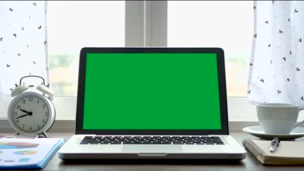 白いカーテンブローで窓の近くの作業オフィステーブルに緑色の画面のクロマキーセットを持つラップトップコンピュータ — ストック動画