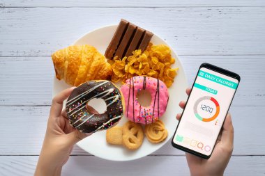 Kalori sayımı ve gıda kontrol konsepti. Akıllı telefonunda elinde donut ve aperatifle Calorie sayaç uygulaması kullanan bir kadın.