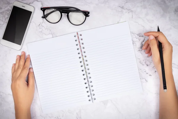 Beyaz mermer masaya boş beyaz bir sayfa defterine el yazısı yazan bir kadın. Gözlükler ve cep telefonu süsleme için masanın üzerinde. — Stok fotoğraf
