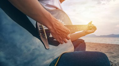 Akustik gitar çalan bir adamın elleri gün batımında kumsalda parmaklarıyla akorları yakalıyor. müzik konsepti çalınıyor
