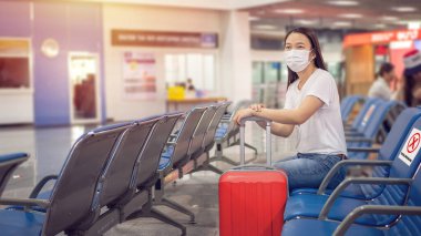 Bagajlı Asyalı turist, hijyenik maske takıyor ve havaalanı terminalinde seyahat sırasında salgını önlemek için sosyal mesafeli sandalyeye oturuyor. Coronavirüs sonrası yeni normallik, covid-19 virüs salgını