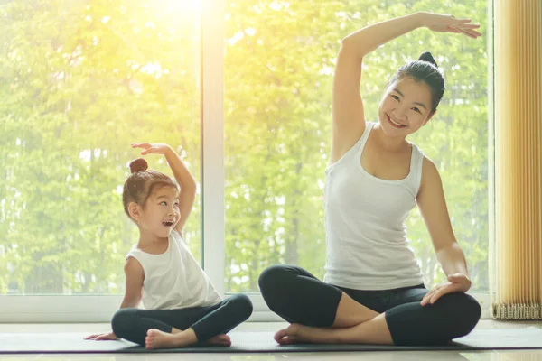 Asian mom practice yoga at home with a adorable daughter sitting next to her, trying to imitate the mother's posture with a smiling face while exercise at home.