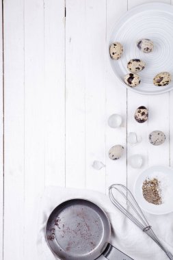 bir tabakta bıldırcın yumurtası, bir kabuk, dayak için bir çırpMa teli ve beyaz ahşap arka plan üzerinde bir tava. yukarıdan görünüm