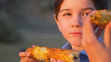anne ve oğlu ızgara mısır yemek, piknikte, nehrin kıyısında, gün batımında, kömürler üzerinde mısır kızartma, 100fps